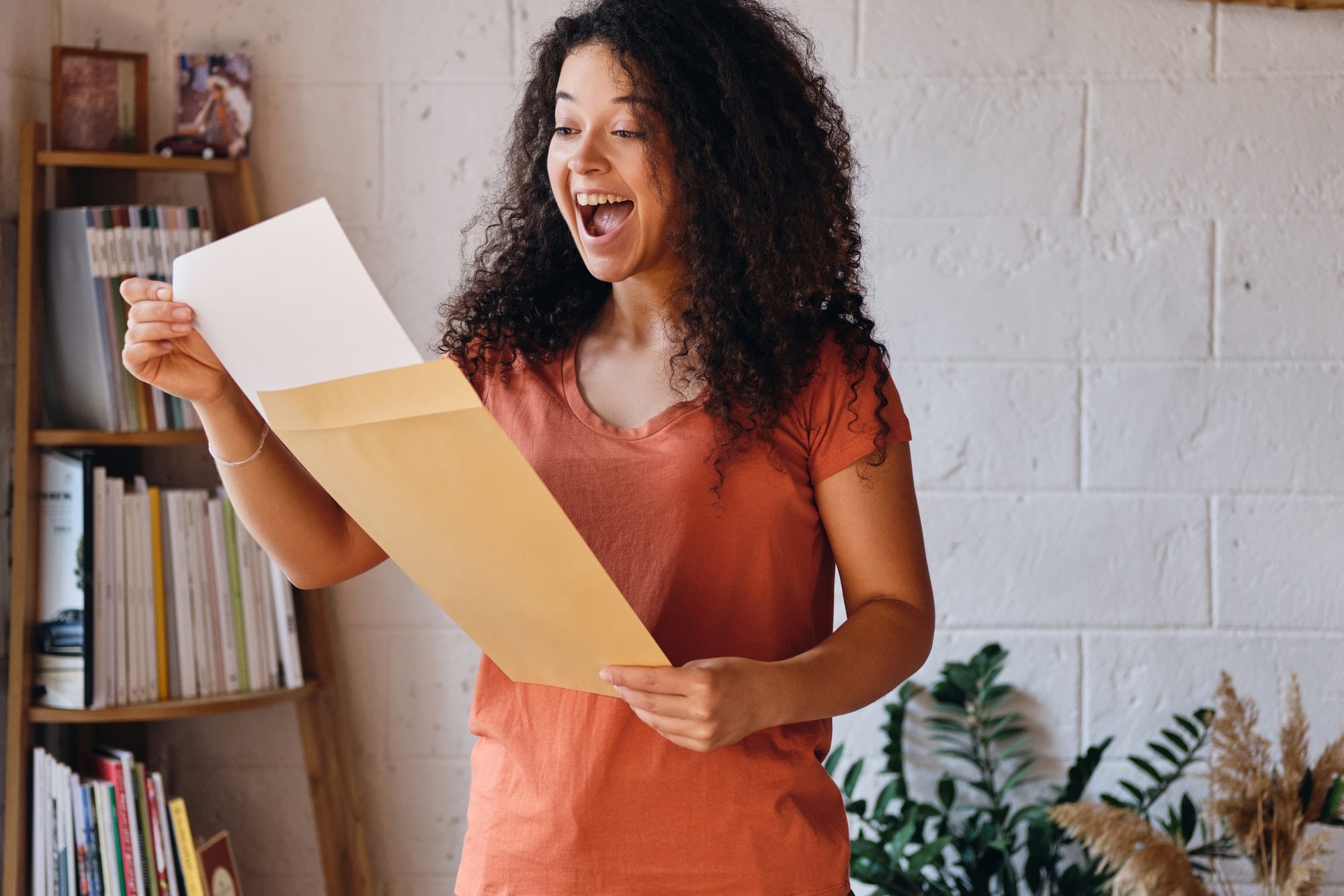 Young joyful woman with dark curly hair in T-shirt happily opening envelope with exam results letter