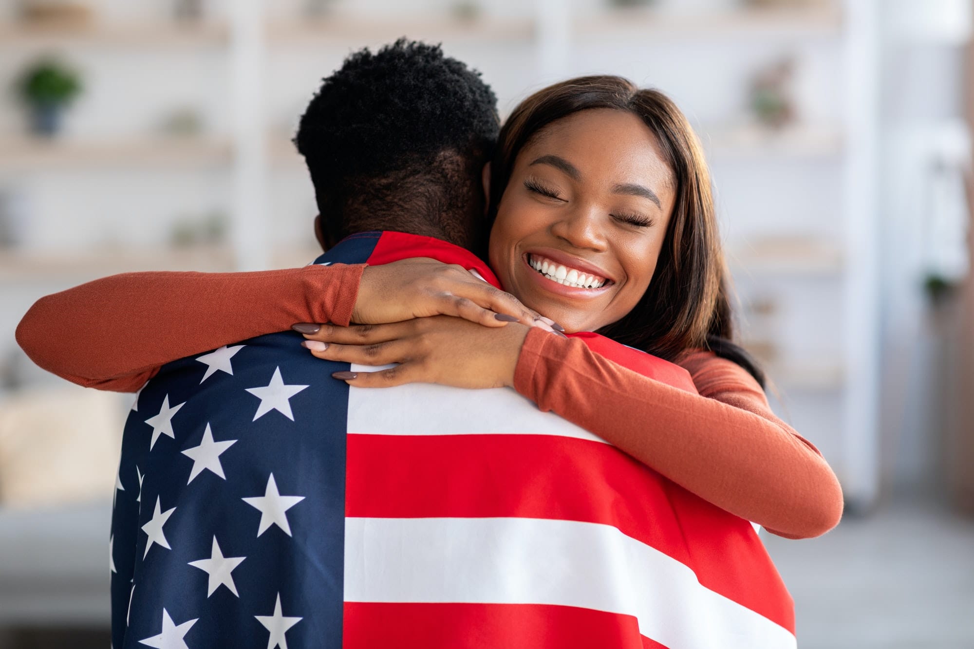 Independence Day. Happy Black Wife Embracing Husband Wrapped In American Flag