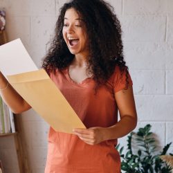 Young joyful woman with dark curly hair in T-shirt happily opening envelope with exam results letter
